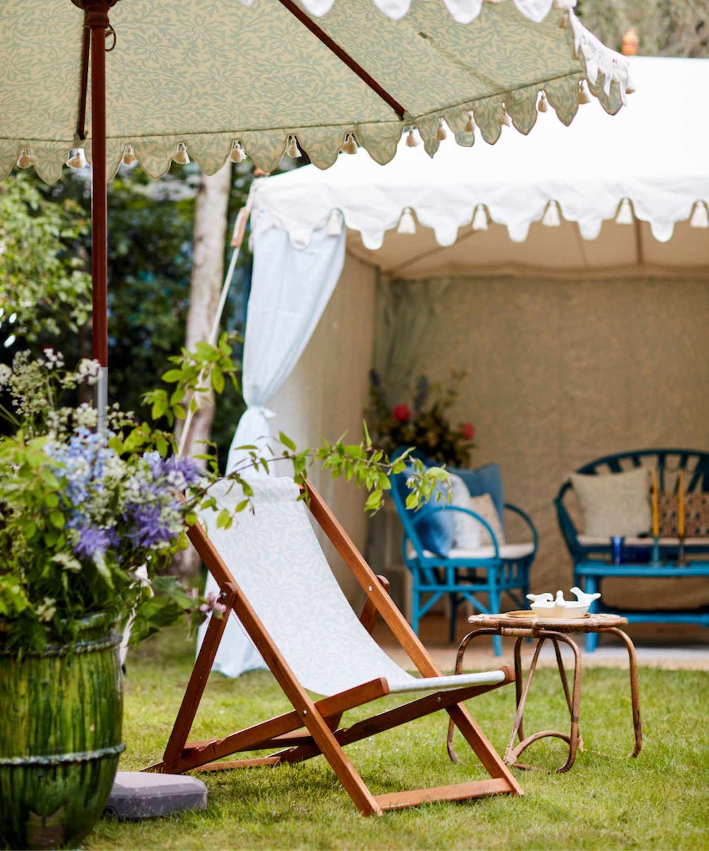 Deckchair and barrel of purple flowers under a parasol with a white tent in the background with blue garden furniture inside