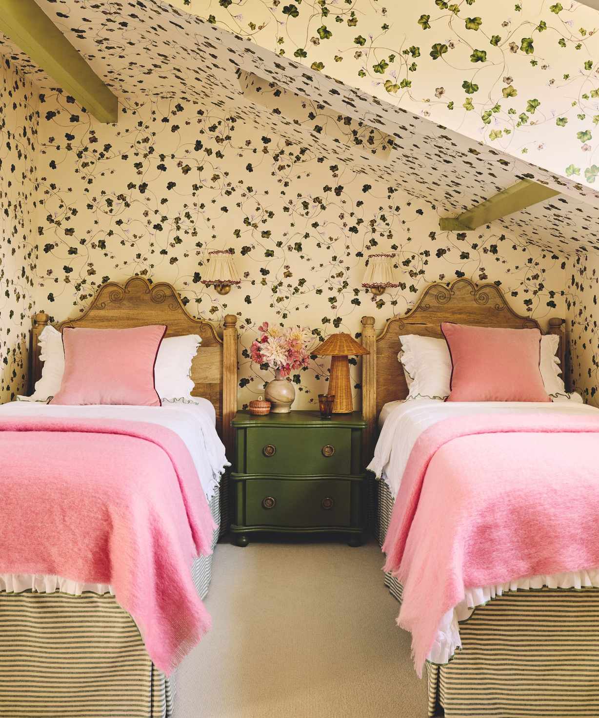 attic twin bedroom witch trailing ivy printed wallpaper on all walls and ceiling with green painted ceiling beams and a green nightstand and pink bed linens