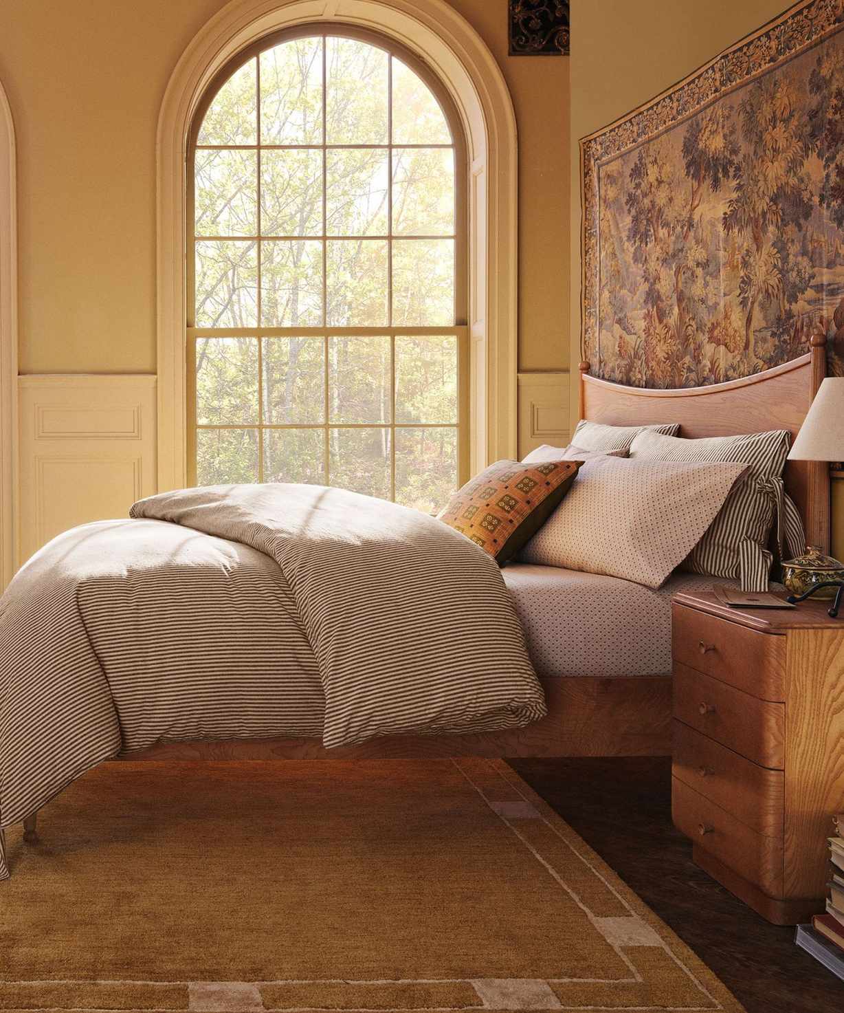 warm butter yellow bedroom with arched windows styled for the Pierce &amp; Ward x West Elm collection with a wooden bed frame, tapestry hung on the wall, and a large ochre rug underneath