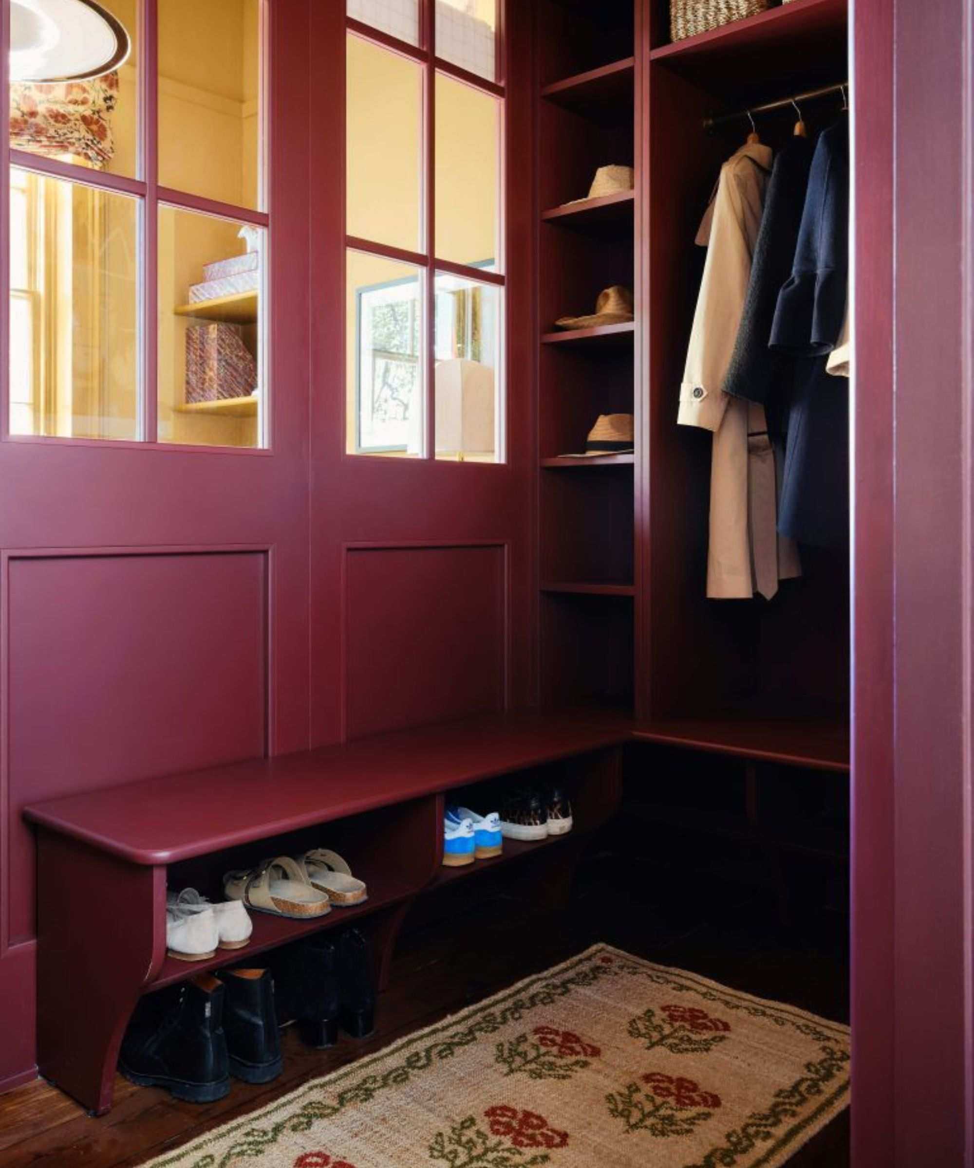 a small boot room in an entryway painted in deep red burgundy paint with a vintage floral rug and storage for coats, shoes, and a bench seat